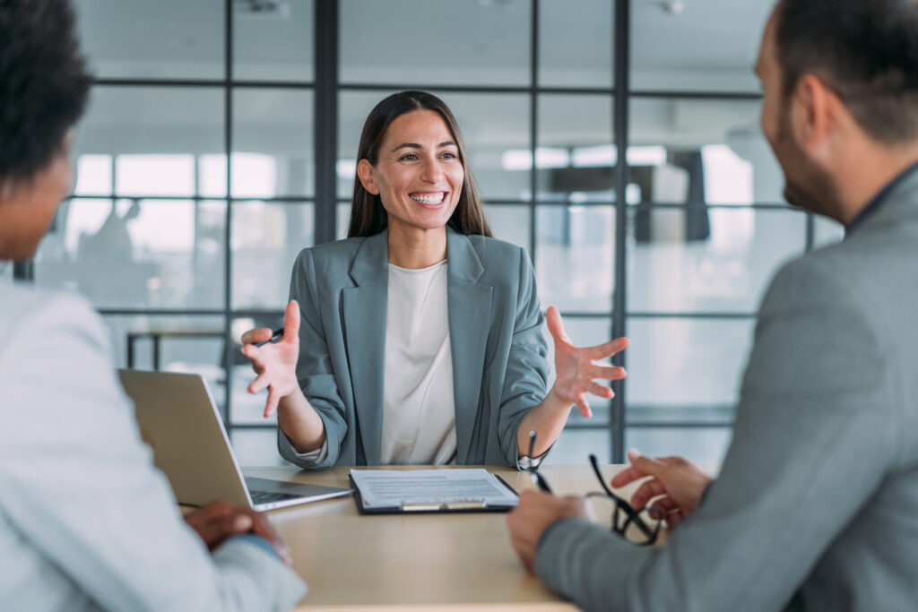A businesswoman smiling and talking with two colleagues at a meeting table in a bright office setting.
