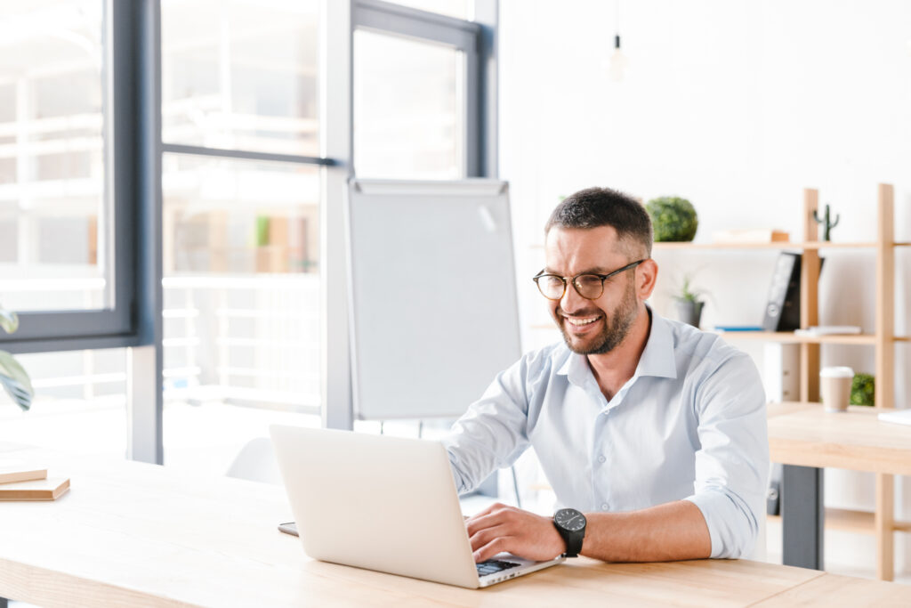 Man in glasses smiling while working on a laptop on public finance in a bright office setting.